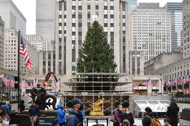 The Rockefeller Christmas Tree, which looks lush with more branches added to it, sits behind some scaffolding in the plaza, with passers-by in the foreground